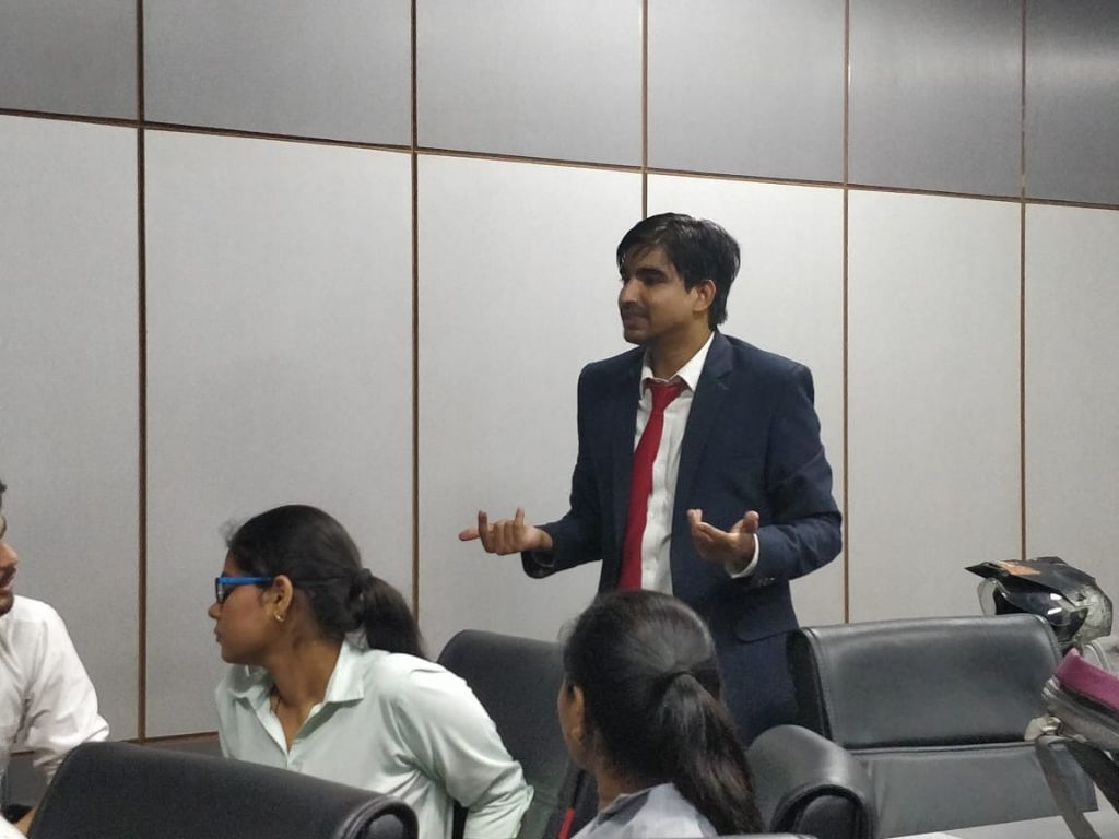 A facilitator in a suit speaking with open hands to students seated around him