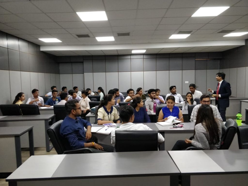 A seminar room with students seated in small groups while a facilitator speaks from the side